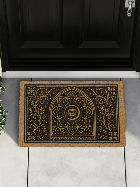 A rectangular, natural brown coir doormat with a non-slip black vinyl backing, viewed from directly above. The mat is made of textured coconut fiber. The design features intricate black line art in a Victorian Gothic and Art Nouveau style. In the center is an All Seeing Eye, or Eye of Providence, enclosed within a radiant starburst and a mandala-like floral pattern. This central motif is set inside an arched frame. The corners and sides are adorned with symmetrical, flowing botanical vines.